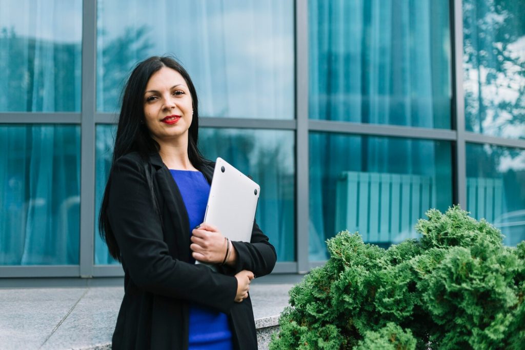 portrait-smiling-businesswoman-holding-laptop-front-glass-building_23-2147970981